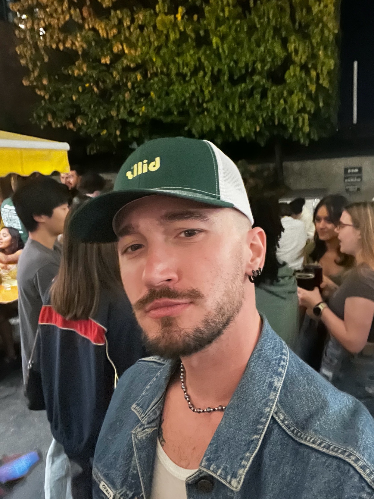 A man takes a selfie at an outdoor event. He wears a denim jacket and a hat.