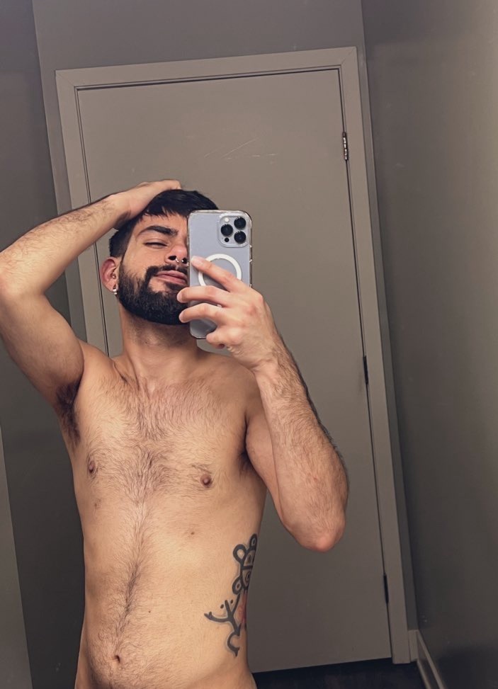 A man takes a mirror selfie, shirtless, in a bathroom. He has dark hair, a beard, and a tattoo on his side.
