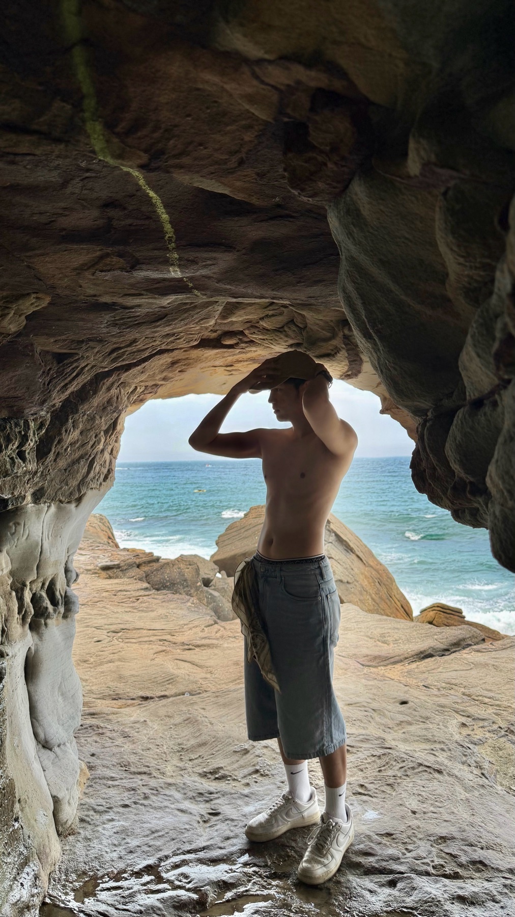 A man stands shirtless in a cave, shielding his eyes from the sun. The ocean and beach are visible in the background.