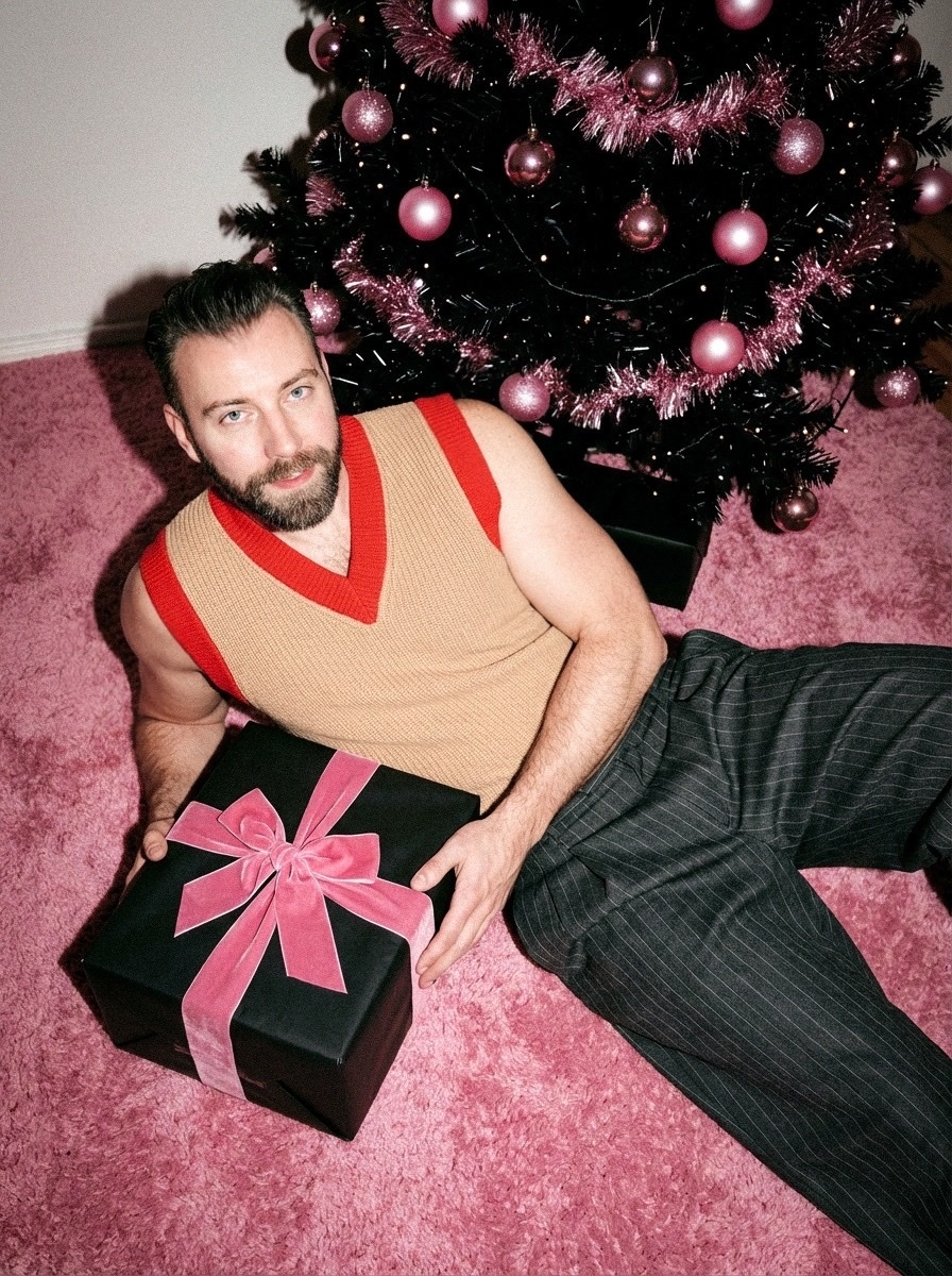 A man poses with a wrapped gift in front of a pink Christmas tree. He is wearing a stylish vest and pants.