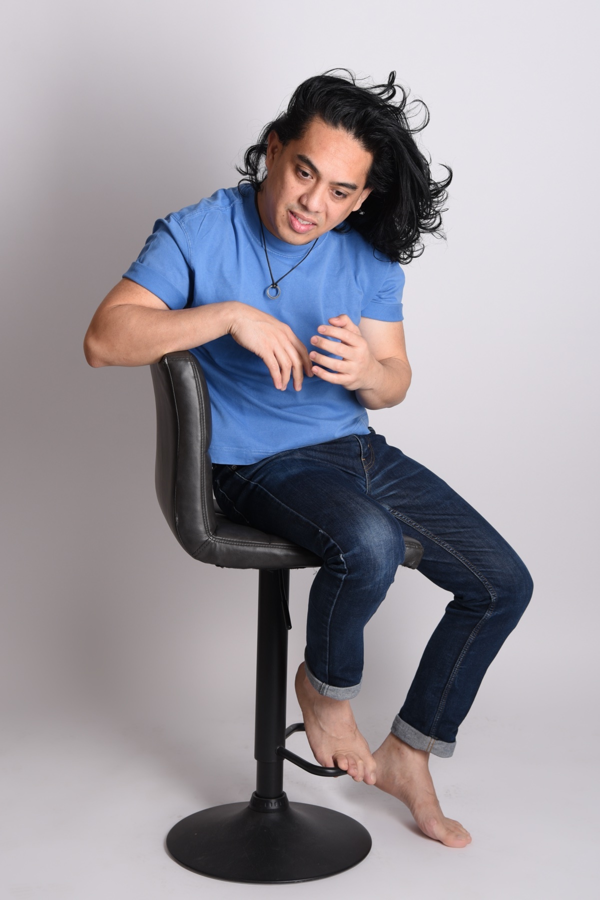 A man with long, dark, wavy hair sits on a bar stool, wearing a blue t-shirt and jeans. He is looking down and to the side, with his hands clasped in front of him.