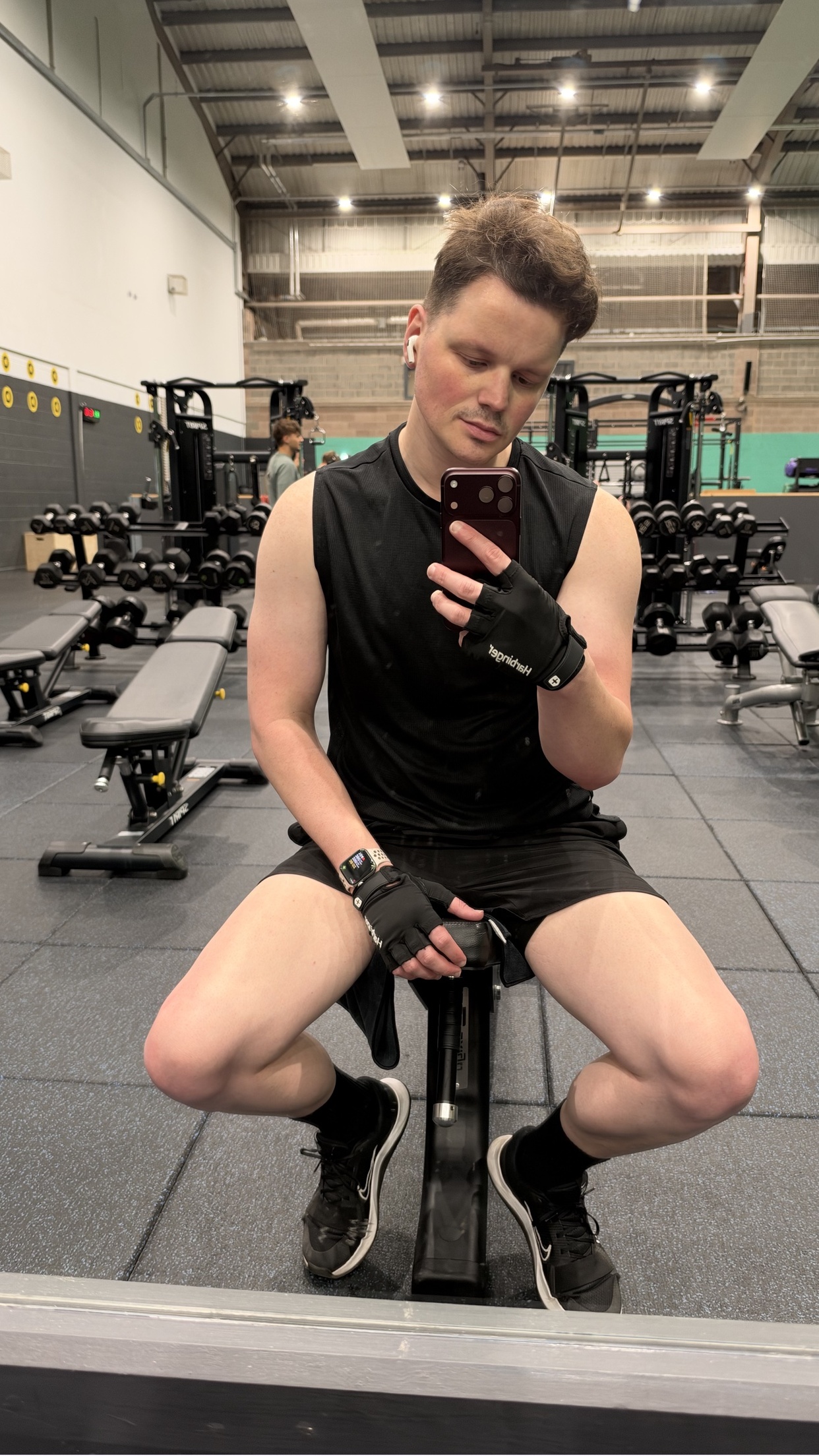 A man takes a selfie in a gym, wearing a black tank top, black shorts, and black athletic gloves. He is sitting on a weight bench with gym equipment in the background.