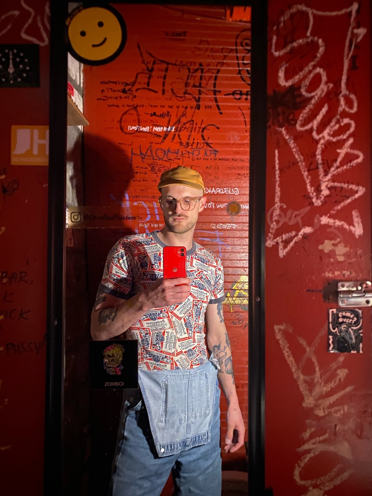 A man takes a selfie in front of a red door covered in graffiti. He is wearing a patterned t-shirt, overalls, and a cap.