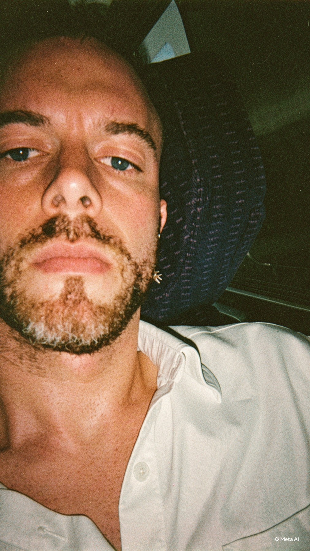 Close-up selfie of a man with stubble and a beard, wearing a white collared shirt. He is looking directly at the camera with a neutral expression.