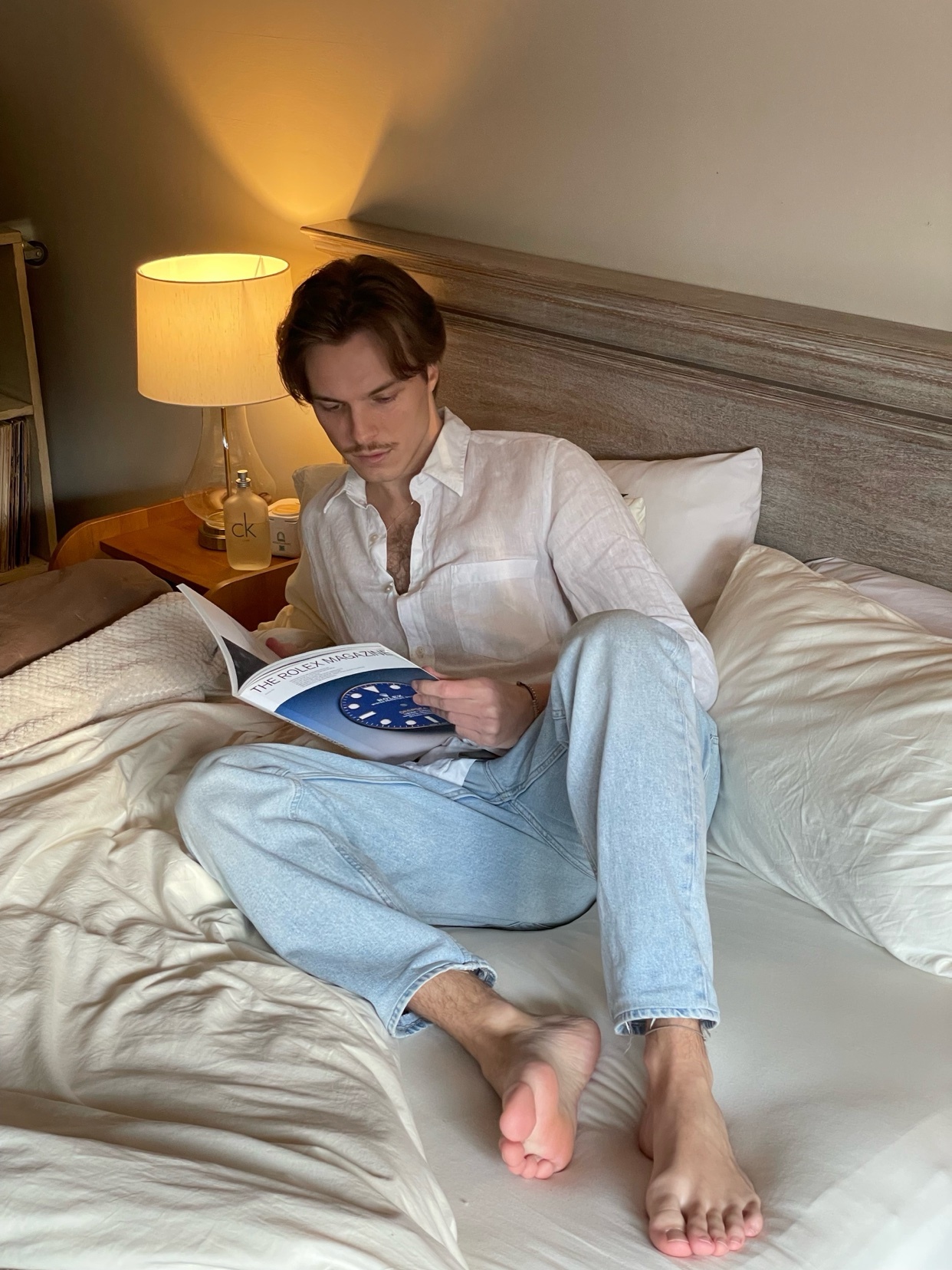 A man with a mustache sits on a bed, wearing a white button-down shirt and light blue jeans, engrossed in reading a magazine.
