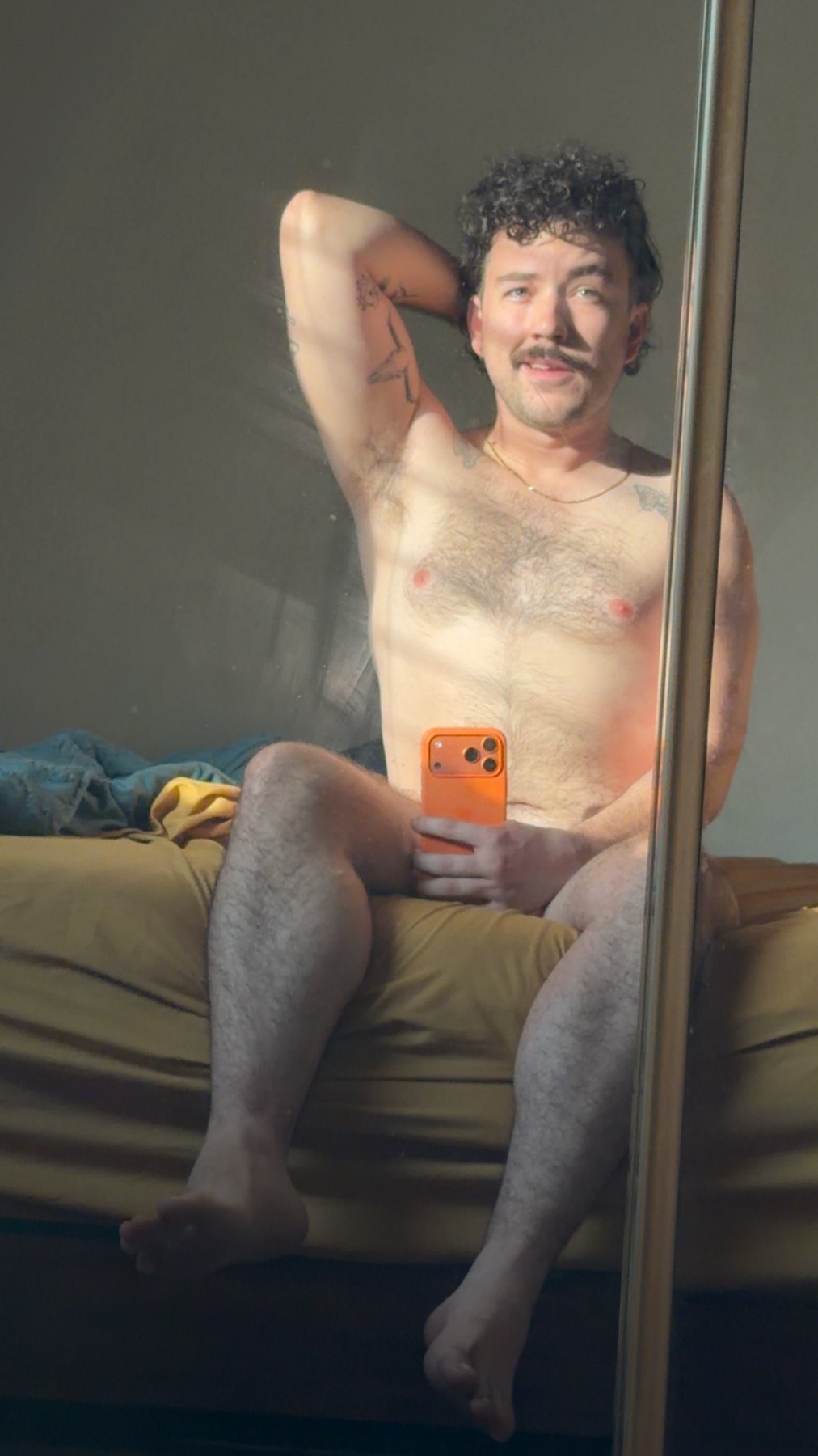 A man takes a shirtless selfie in front of a mirror. The photo is taken in a bedroom with sunlight streaming in.