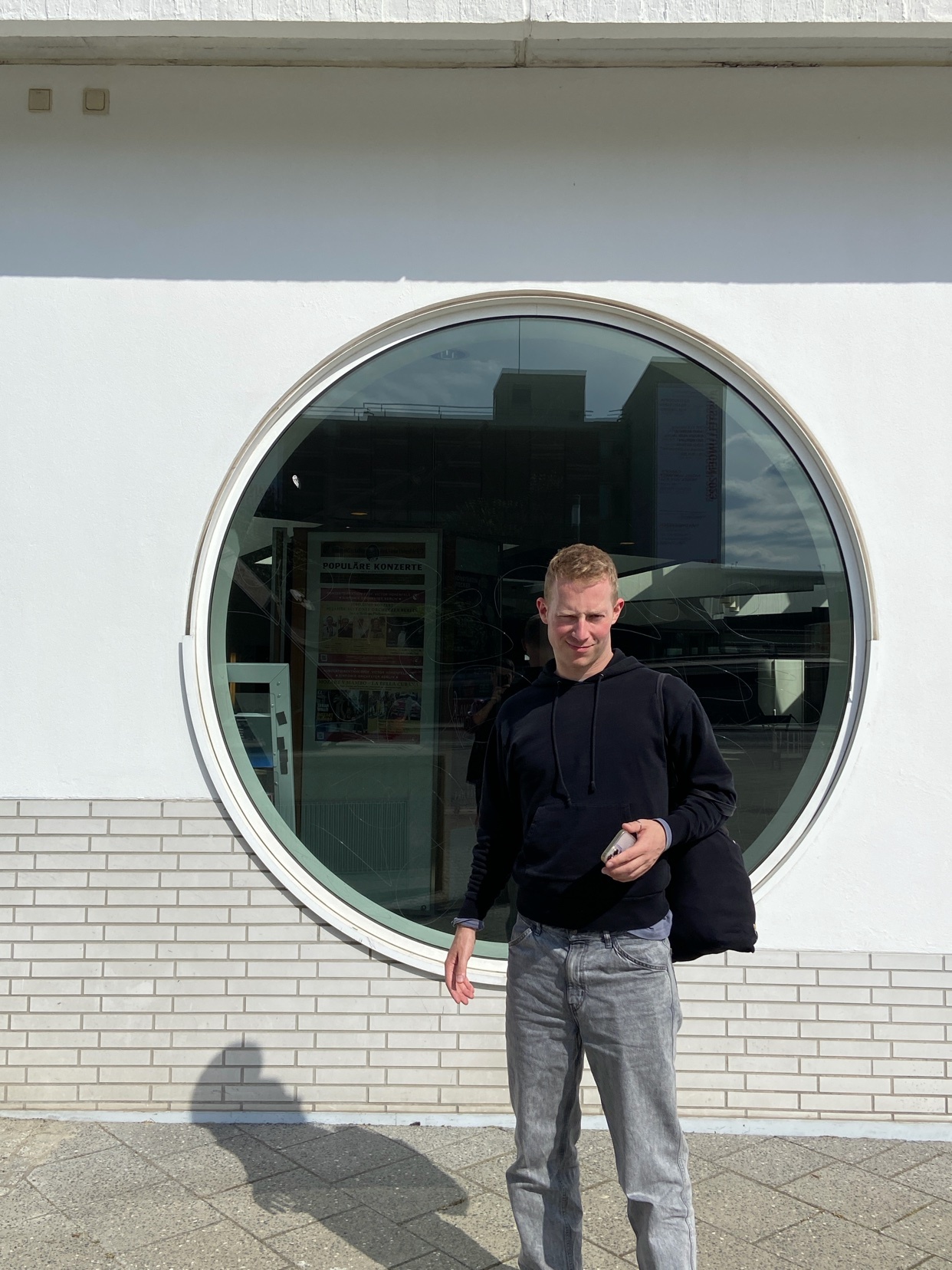 A man in a black hoodie and grey jeans stands in front of a building with a large, circular window. He is holding a phone in his right hand and a bag is slung over his left shoulder.