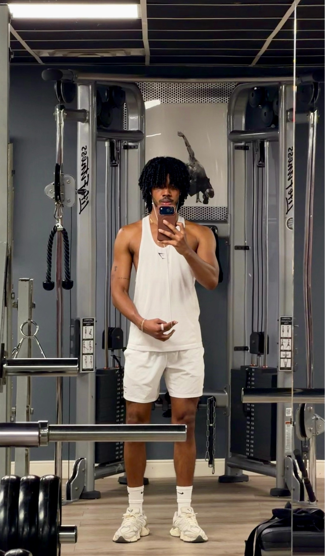 A man takes a mirror selfie in a gym, wearing a white tank top and shorts. He has curly dark hair and is standing in front of weight machines.