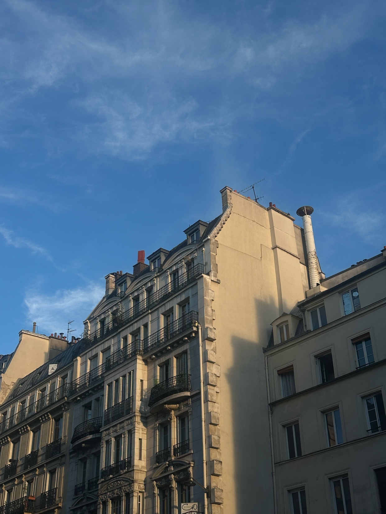A low-angle shot of Parisian buildings against a bright blue sky with wispy clouds. The architecture features classic European design with balconies and mansard roofs.