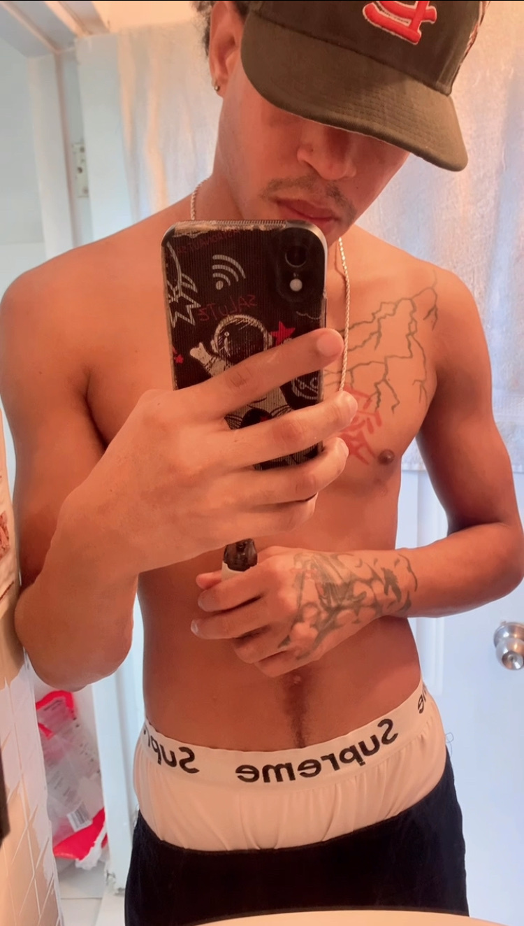 A young man with tattoos on his arms and chest takes a selfie in a bathroom. He is wearing a baseball cap and underwear, and his torso is bare.