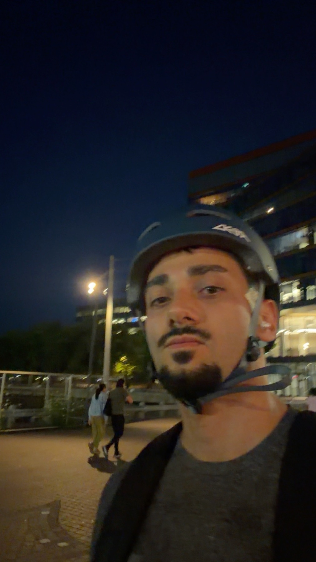 A man wearing a helmet and a grey t-shirt poses for a selfie at night while rollerblading. In the background, a building and a few people can be seen.