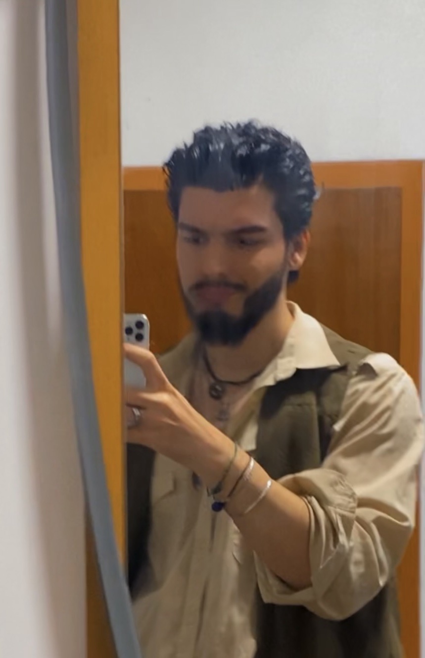 A man with dark, curly hair and a beard takes a selfie in a mirror. He is wearing a vest over a button-up shirt and several pieces of jewelry.