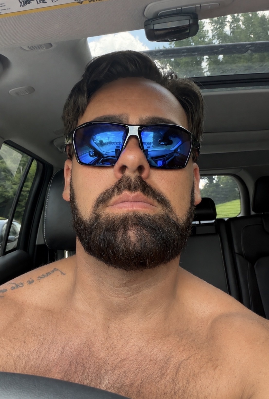 A man with a beard and sunglasses is looking at the camera while sitting in a car. The sun is shining, and there are trees in the background.