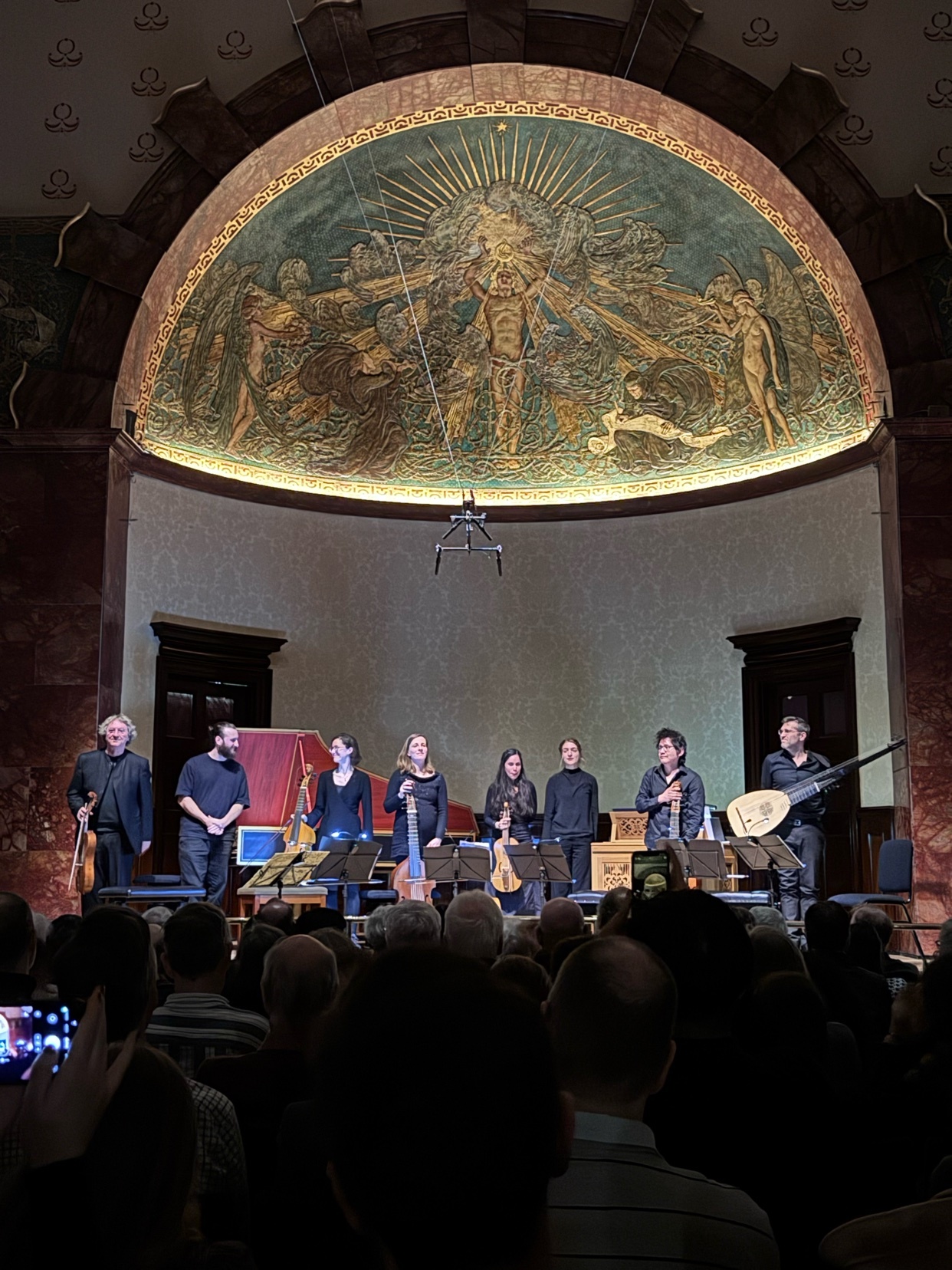 A group of musicians performs on a stage in front of an ornate mural. The musicians are playing various string instruments, including a lute and violins. An audience is visible in the foreground, seated in the dark.