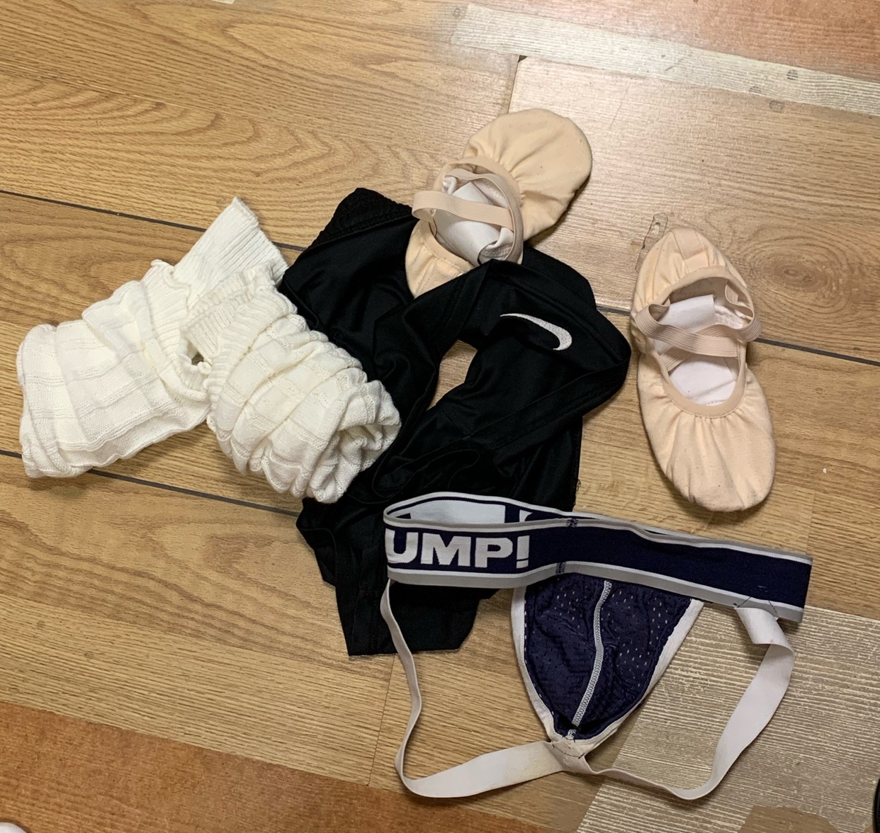 A collection of ballet shoes, jockstrap, and other workout clothing on a wooden floor. The items suggest a completed ballet practice session.