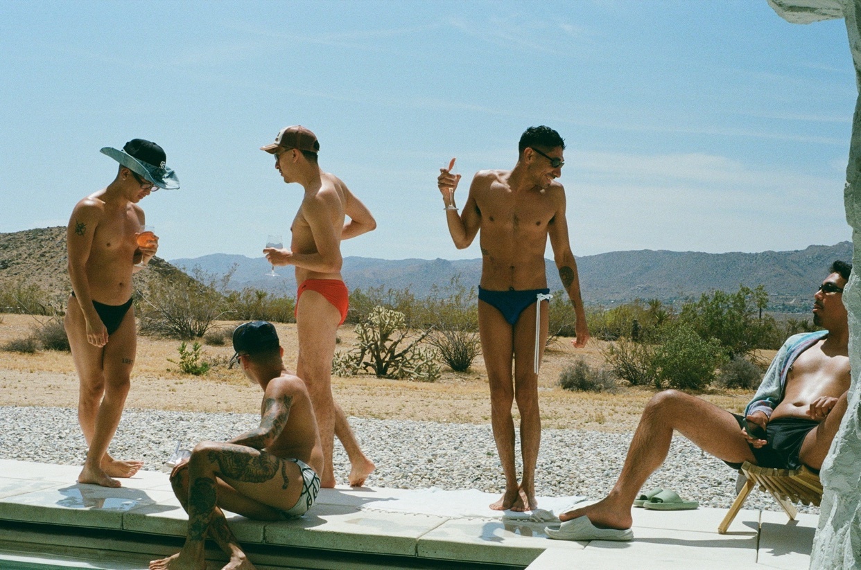 A group of five men in swimwear gather by a pool in a desert landscape. The men are dressed in various swim trunks and some are wearing hats and sunglasses.