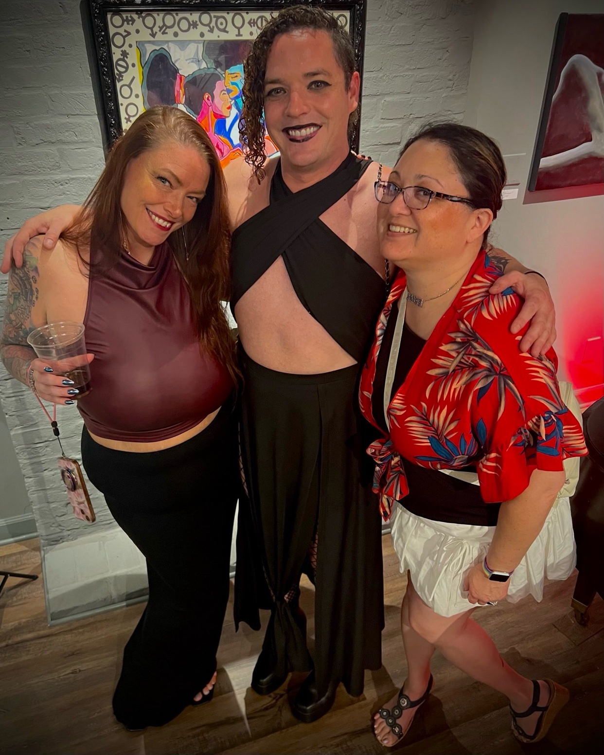 Three people pose for a photo at an erotic art expo. The person in the center, who appears non-binary, wears a black halter top and skirt. To their left, a woman in a maroon top and black skirt holds a drink. To their right, another woman in a red fl