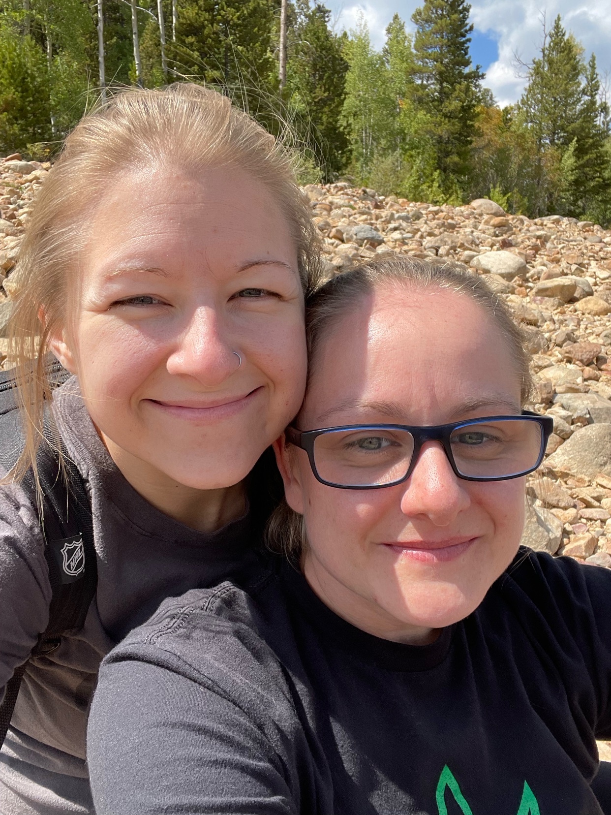 A close-up selfie of two smiling women outdoors with a rocky shoreline and trees in the background.