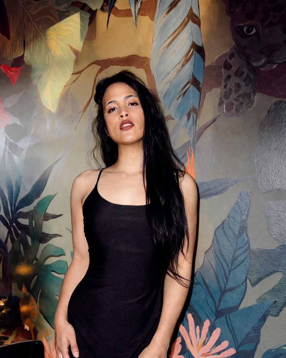 A woman with long, dark hair poses in front of a vibrant tropical-themed mural. She is wearing a simple black spaghetti strap dress.
