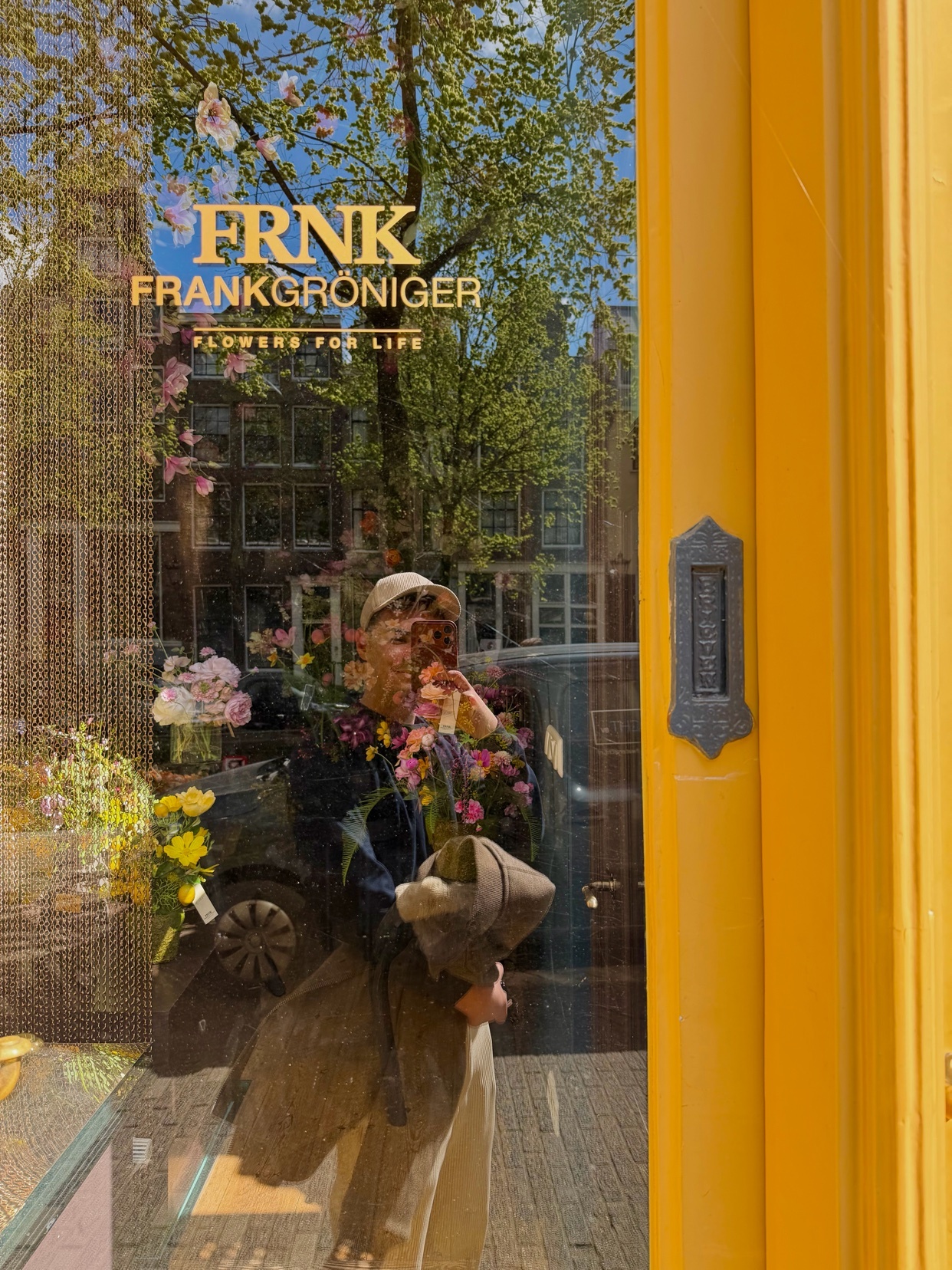 A reflection in a shop window shows a person holding a bouquet of flowers in front of a florist shop. The shop's name, FRNK FRANKGRONIGER FLOWERS FOR LIFE, is visible on the glass.