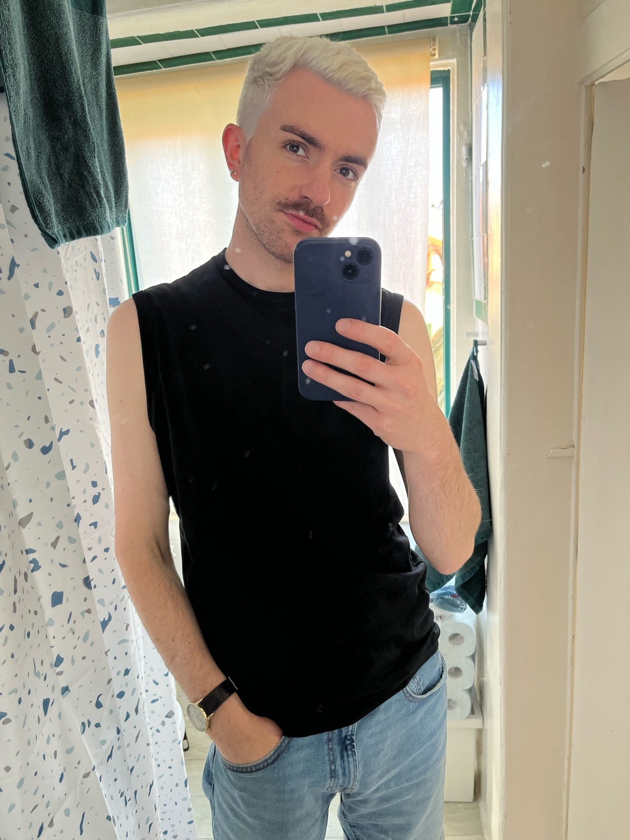 A man with blonde hair and a mustache takes a selfie in a bathroom mirror. He is wearing a black sleeveless shirt and blue jeans.