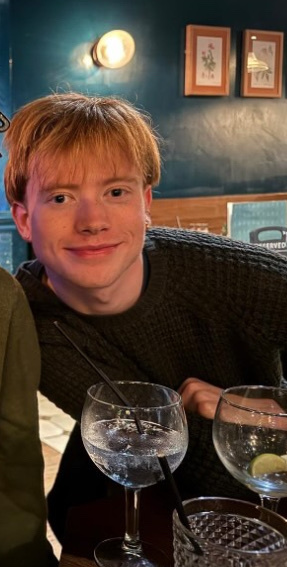 A young man with ginger hair smiles at the camera. He is wearing a sweater and appears to be at a party.