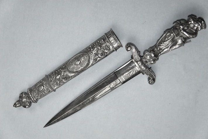 An ornate silver dagger with a decorative handle and a matching sheath rests on a plain background. The dagger features intricate carvings on its handle and sheath, suggesting it is an antique or decorative piece.