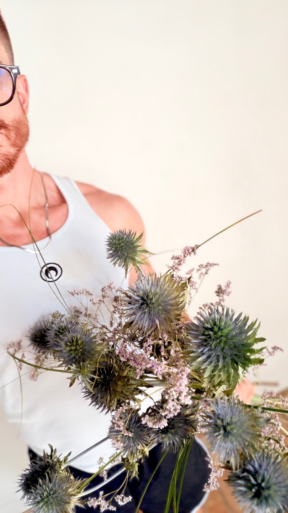 A man wearing glasses and a white tank top holds a bouquet of thistle and dried flowers. He has a beard and stubble, and is wearing a necklace.
