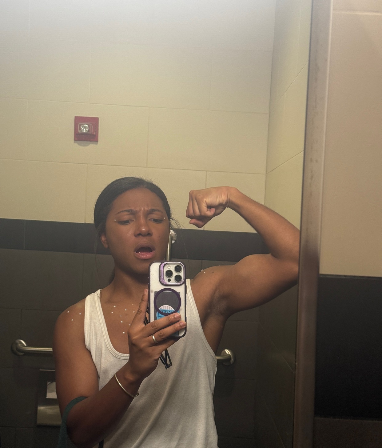 A woman in a white tank top flexes her bicep while taking a selfie in a bathroom mirror. She has a surprised expression on her face and is wearing a bracelet on her left wrist.
