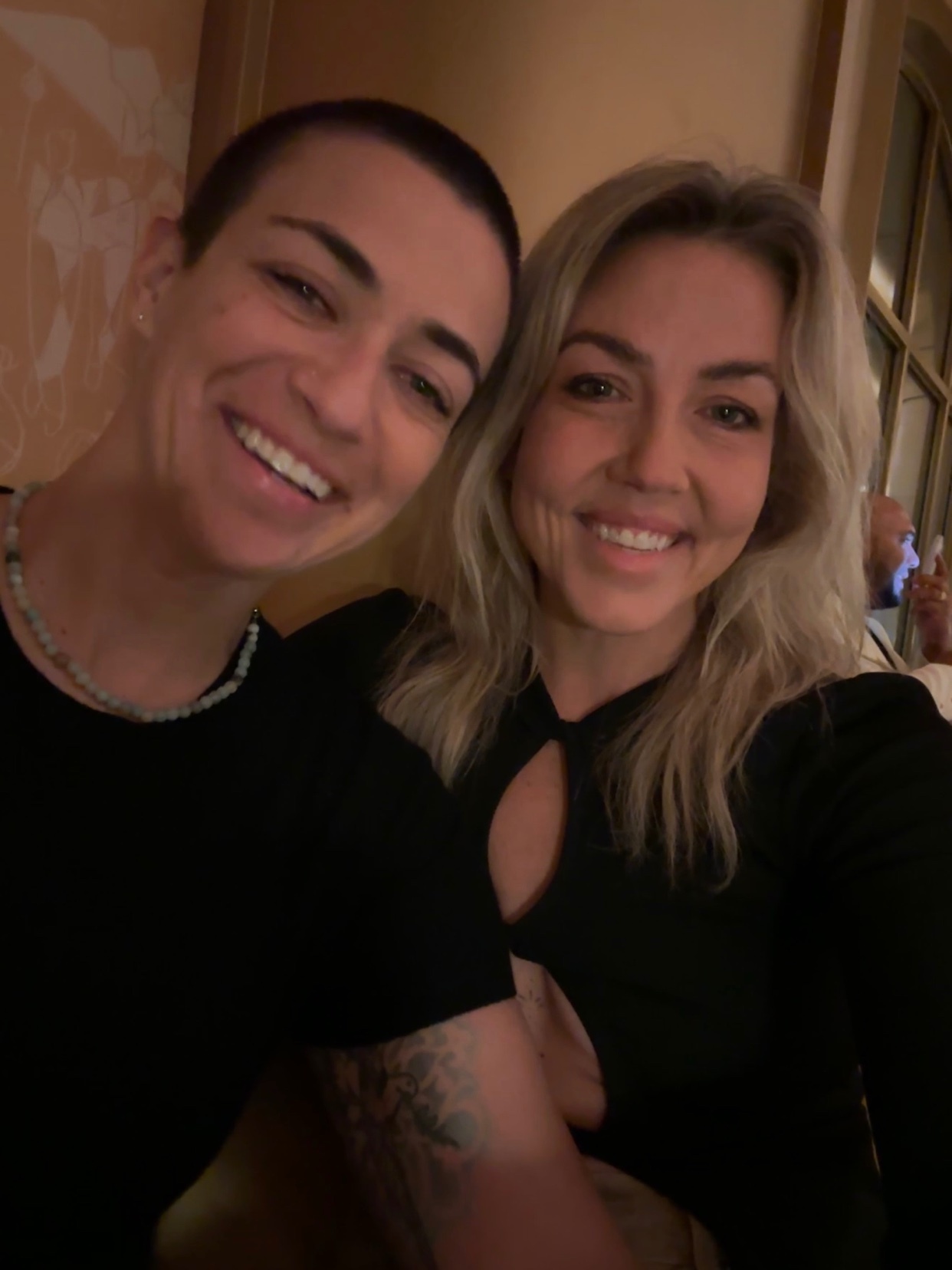A close-up selfie shows two smiling women. The woman on the left has short, dark hair and is wearing a black shirt and a beaded necklace. The woman on the right has blonde hair and is wearing a black top with a cutout detail.