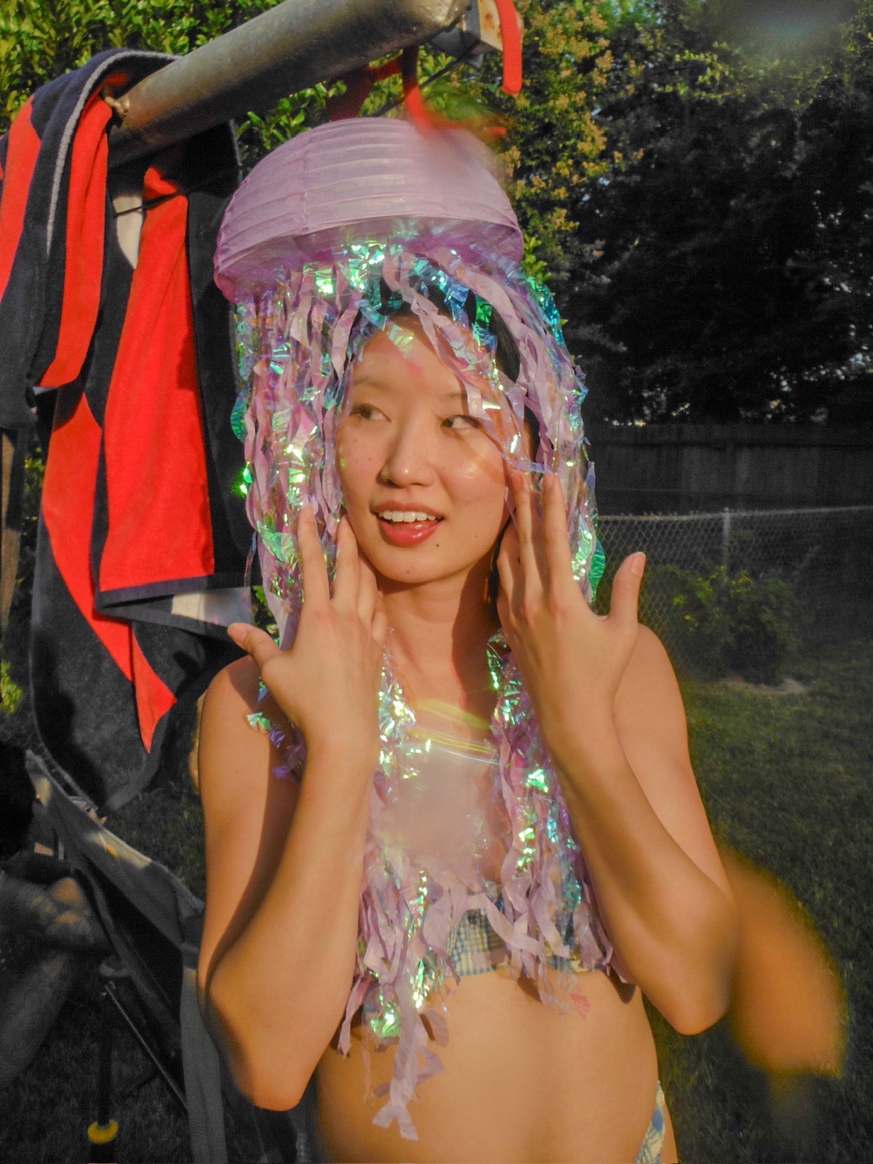 A woman poses outdoors wearing a whimsical, iridescent jellyfish costume. The costume features a purple lantern headpiece with shimmering, dangling tentacles.
