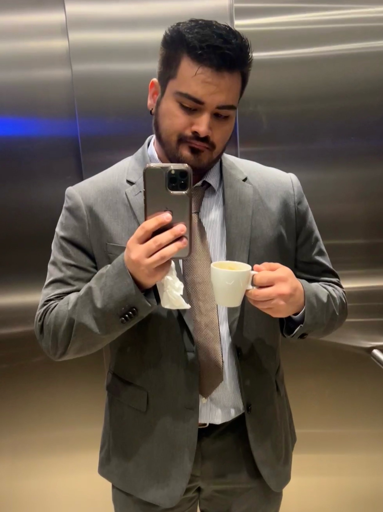A man in a suit and tie takes a selfie in an elevator while holding a cup of coffee and a tissue.