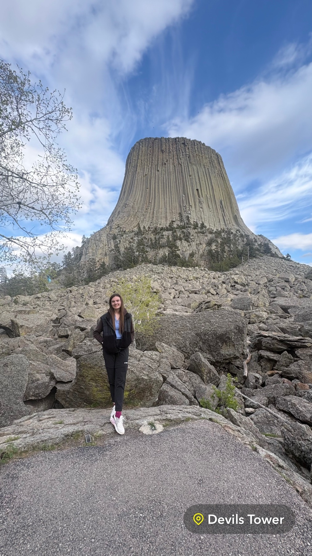 A woman stands in front of the iconic Devils Tower National Monument in Wyoming. The massive rock formation dominates the background under a bright blue sky with scattered clouds.