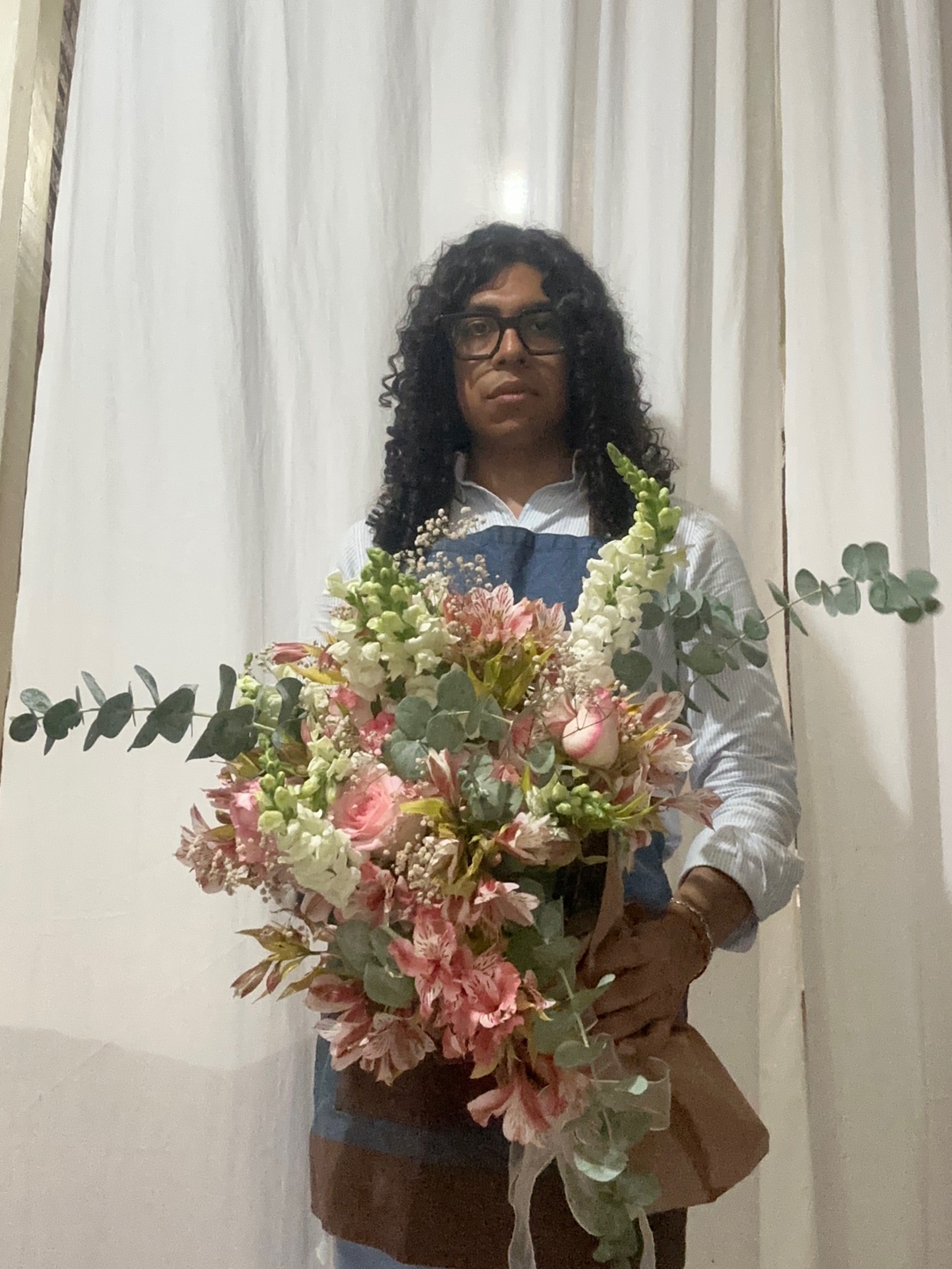 A man with curly dark hair and glasses stands holding a large bouquet of pink and white flowers. He is wearing a light blue shirt and a denim apron.