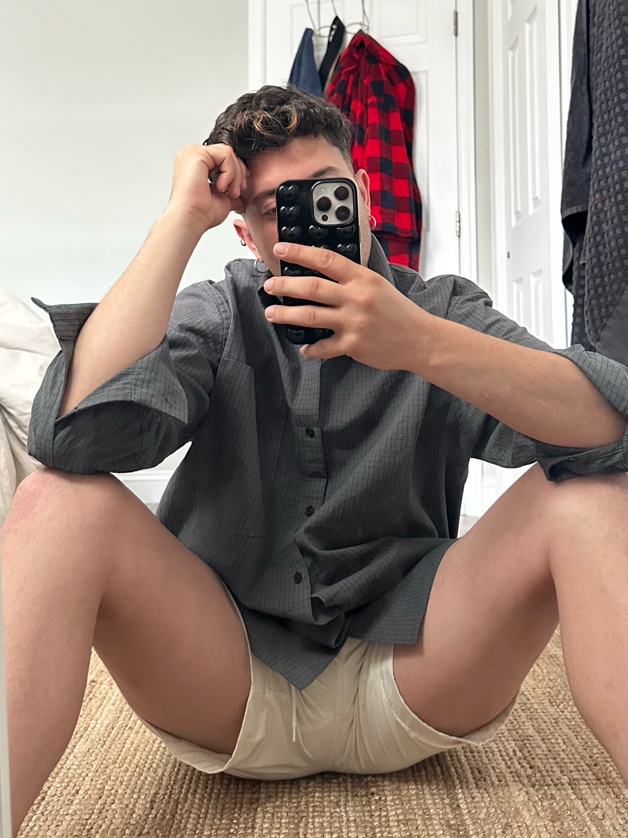 A man takes a mirror selfie, seated on a rug. He wears a grey button-down shirt and white shorts, showcasing a modern and casual style.
