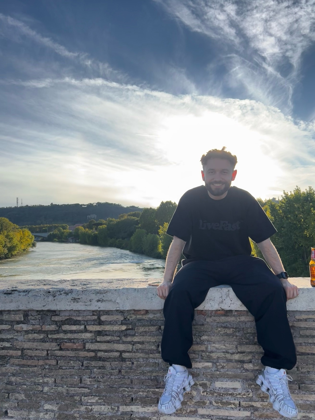 A man sits on a brick wall overlooking a river and cityscape. The sky is blue with some clouds.