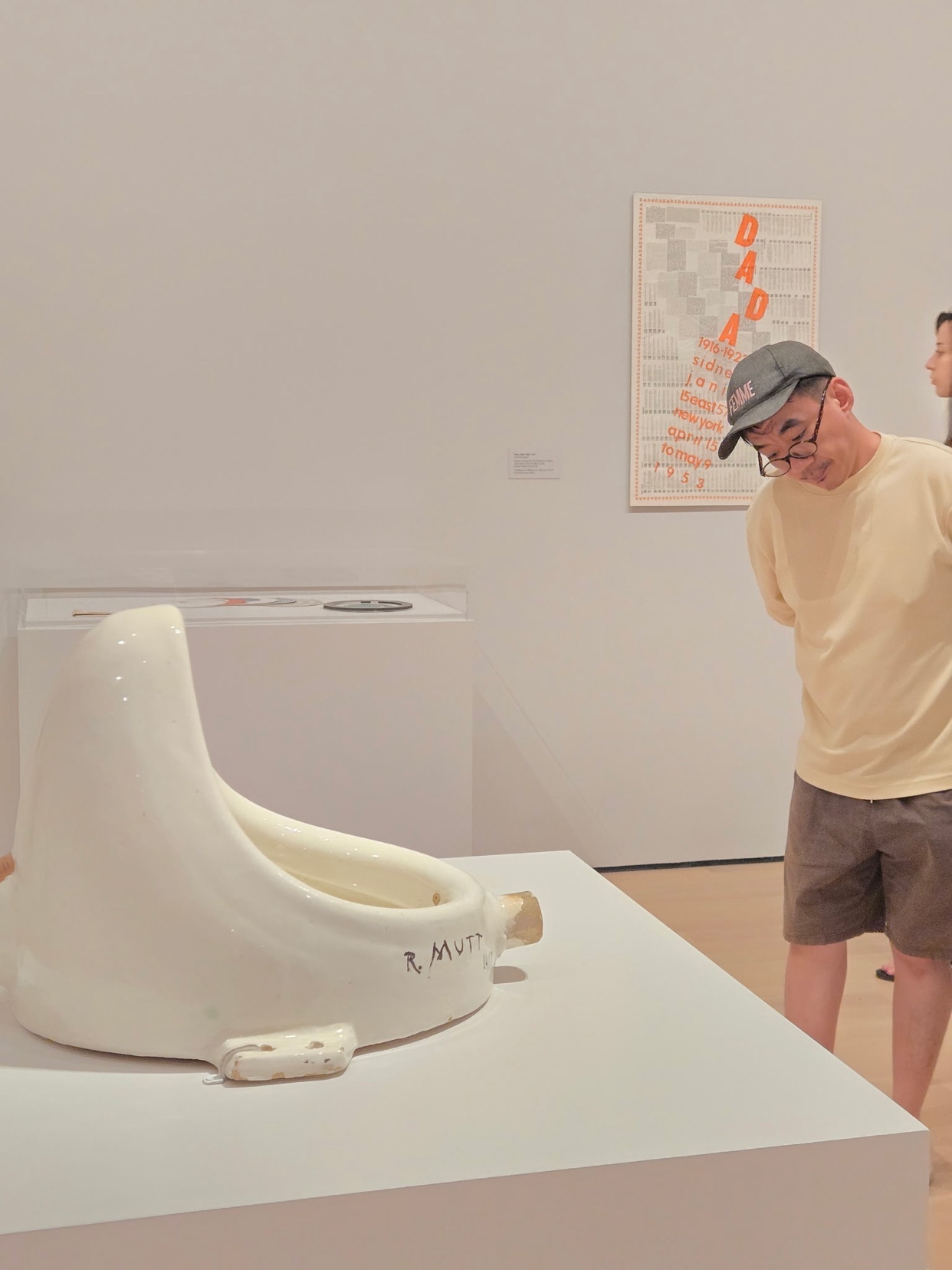 A man wearing glasses and a baseball cap looks at a white porcelain urinal sculpture displayed on a pedestal in an art gallery. Behind the sculpture, a poster with the word "DADA" in large orange letters is visible on the wall.