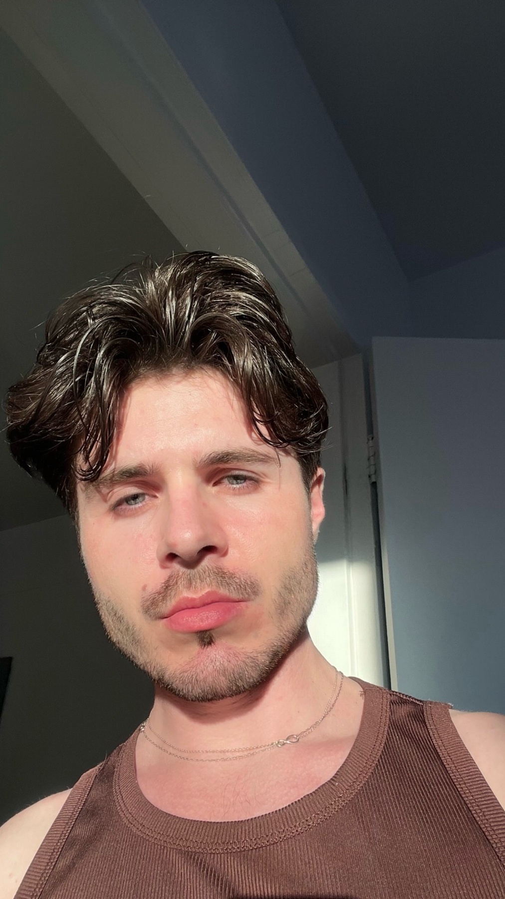 A man poses for a selfie, showcasing his features. He is shirtless, with dark hair and a brown tank top.