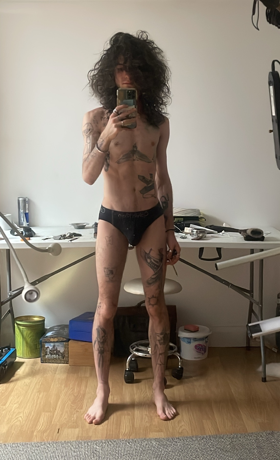 A shirtless man poses for a mirror selfie, showcasing his tattoos and physique. The photo is taken in a modern bedroom setting.