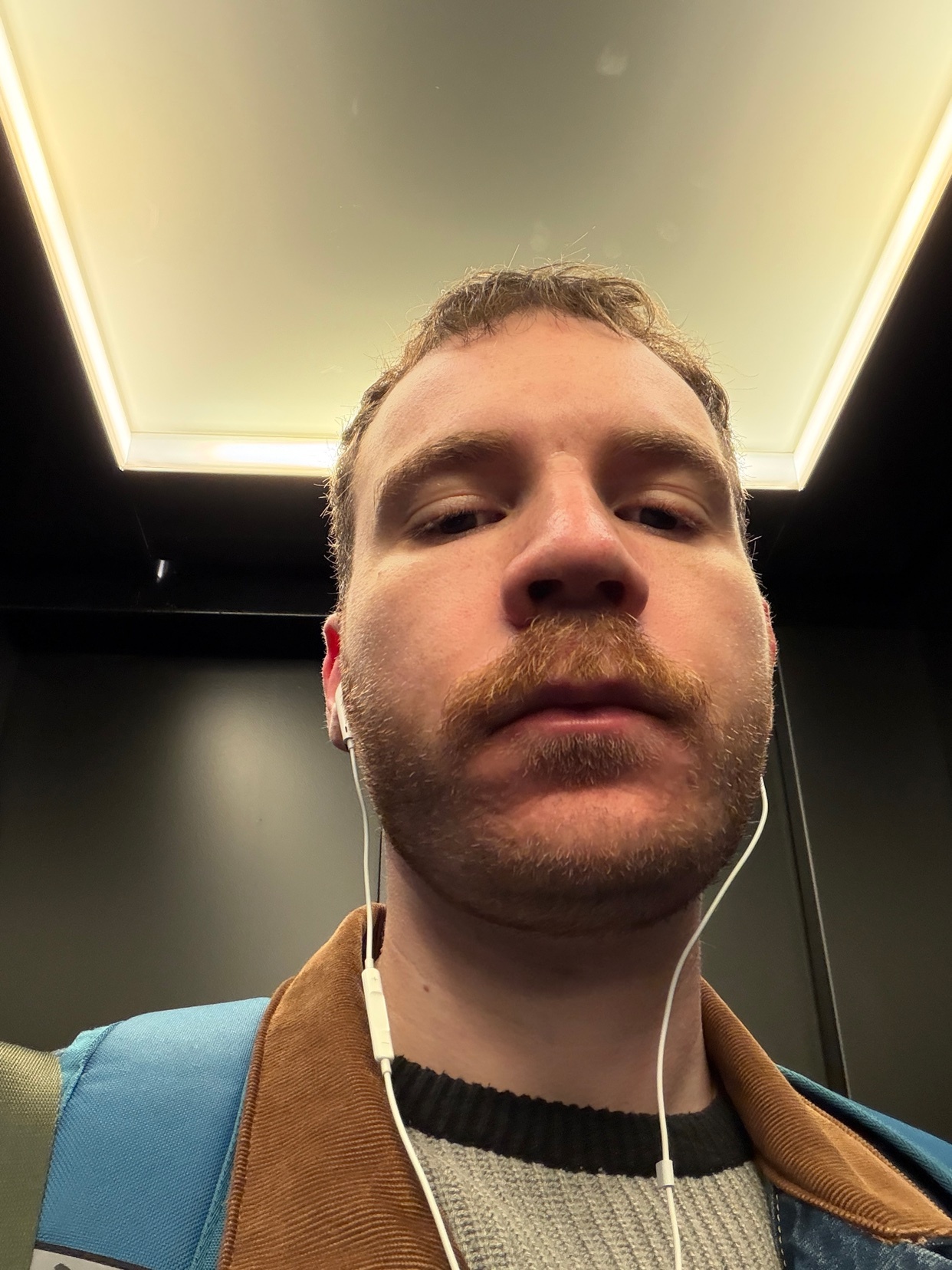 A man with a mustache and beard wears earbuds and a brown jacket over a gray sweater. He appears tired after work.