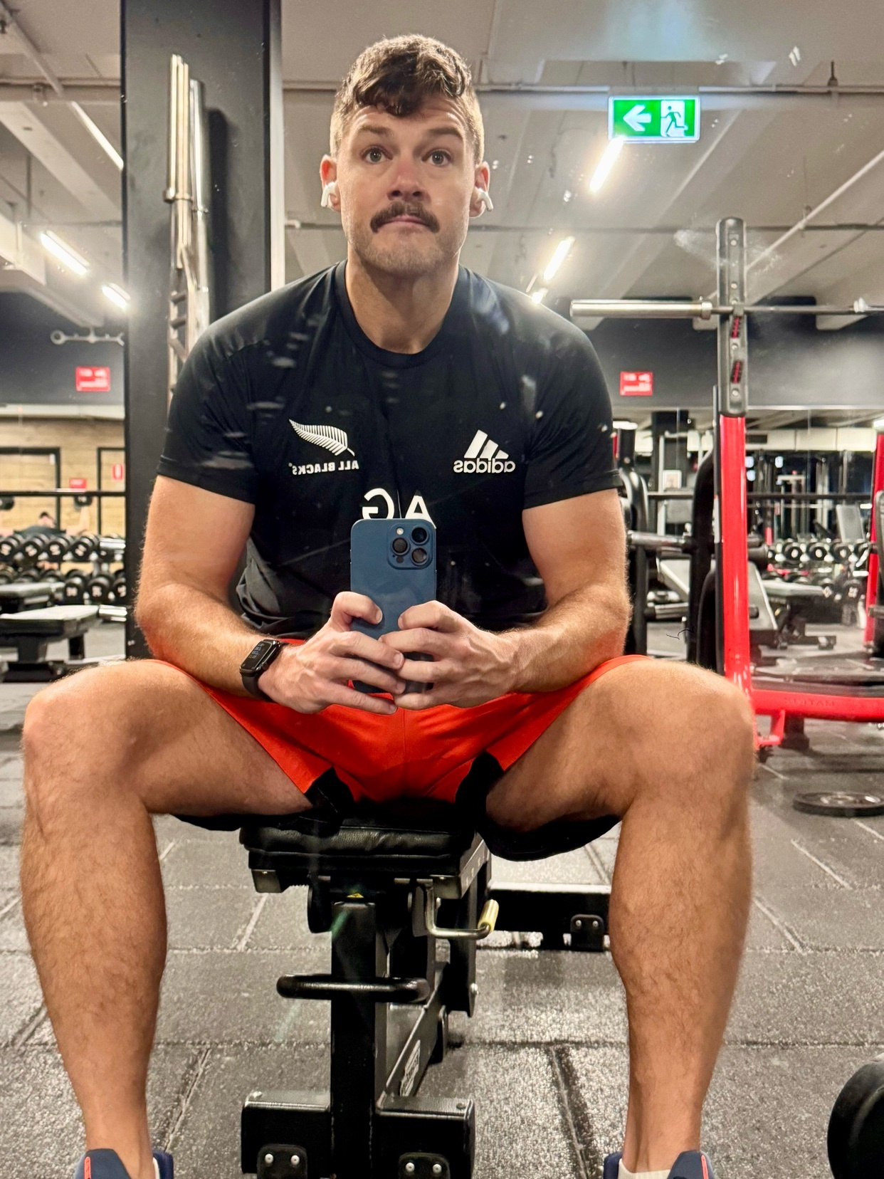 A man with a prominent mustache sits on a weight bench in a gym, holding his phone to take a selfie. He is wearing a black t-shirt and orange shorts, and appears to be in the middle of a workout session.