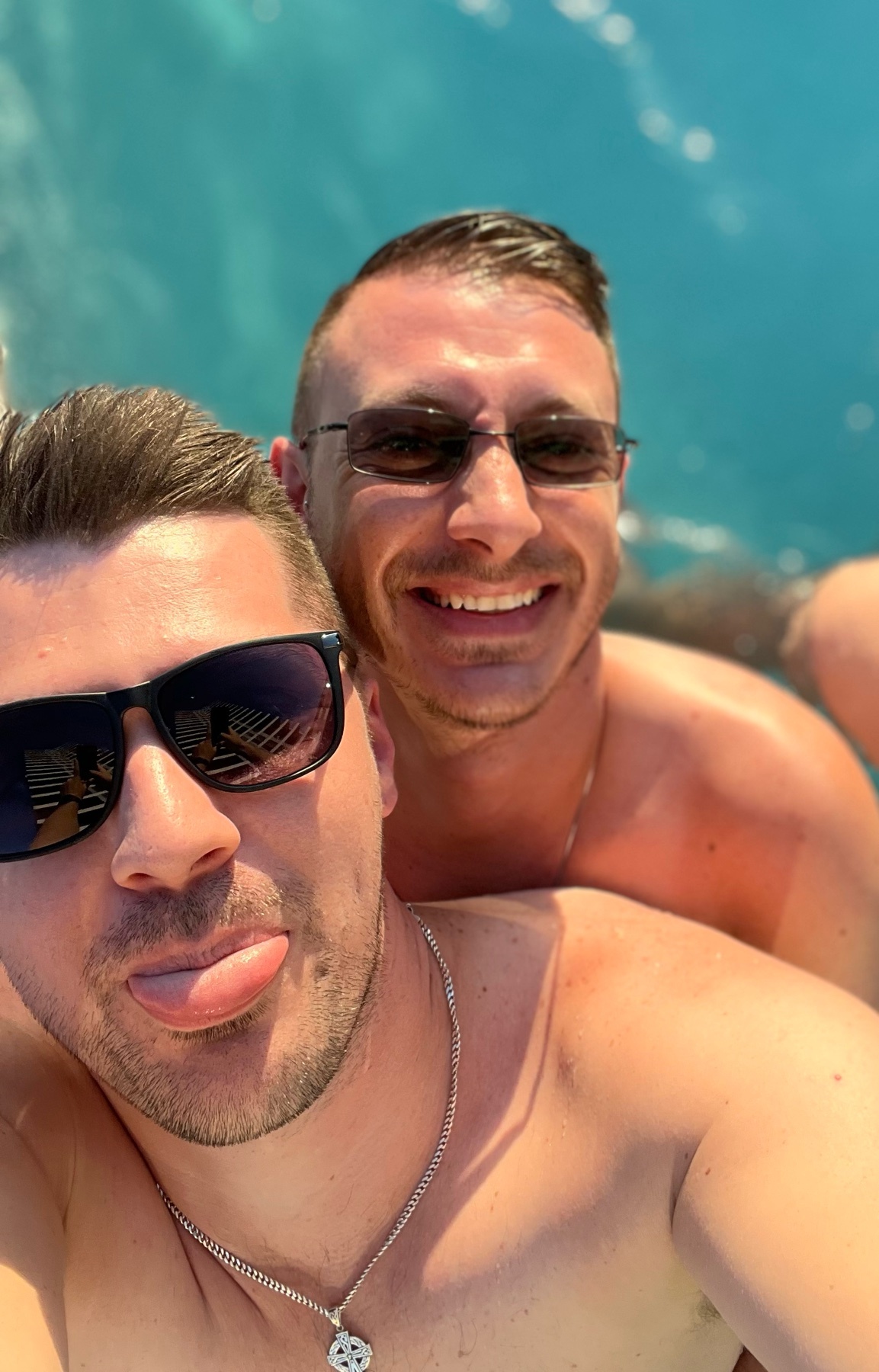 A close-up selfie of two men in sunglasses, one sticking his tongue out, in a pool on a sunny day.