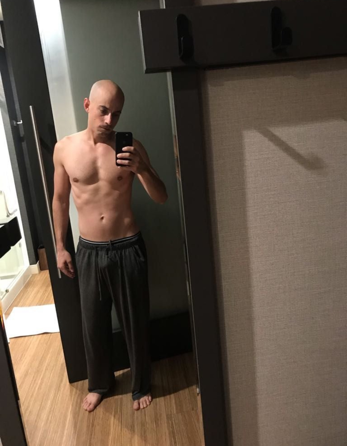 A man takes a shirtless mirror selfie, showcasing his physique. The photo highlights his muscular build and bald head.