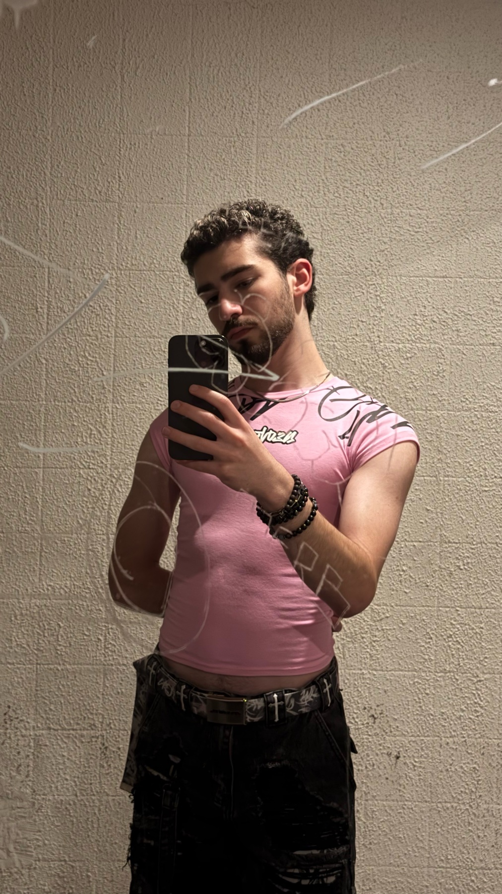 A man takes a selfie in a pink, fitted t-shirt and dark pants. He has curly dark hair and stubble, and is wearing beaded bracelets.