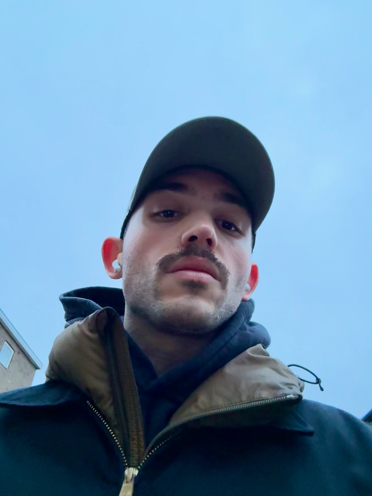 A man takes a selfie, wearing a cap and a jacket. The man has a mustache and dark hair.