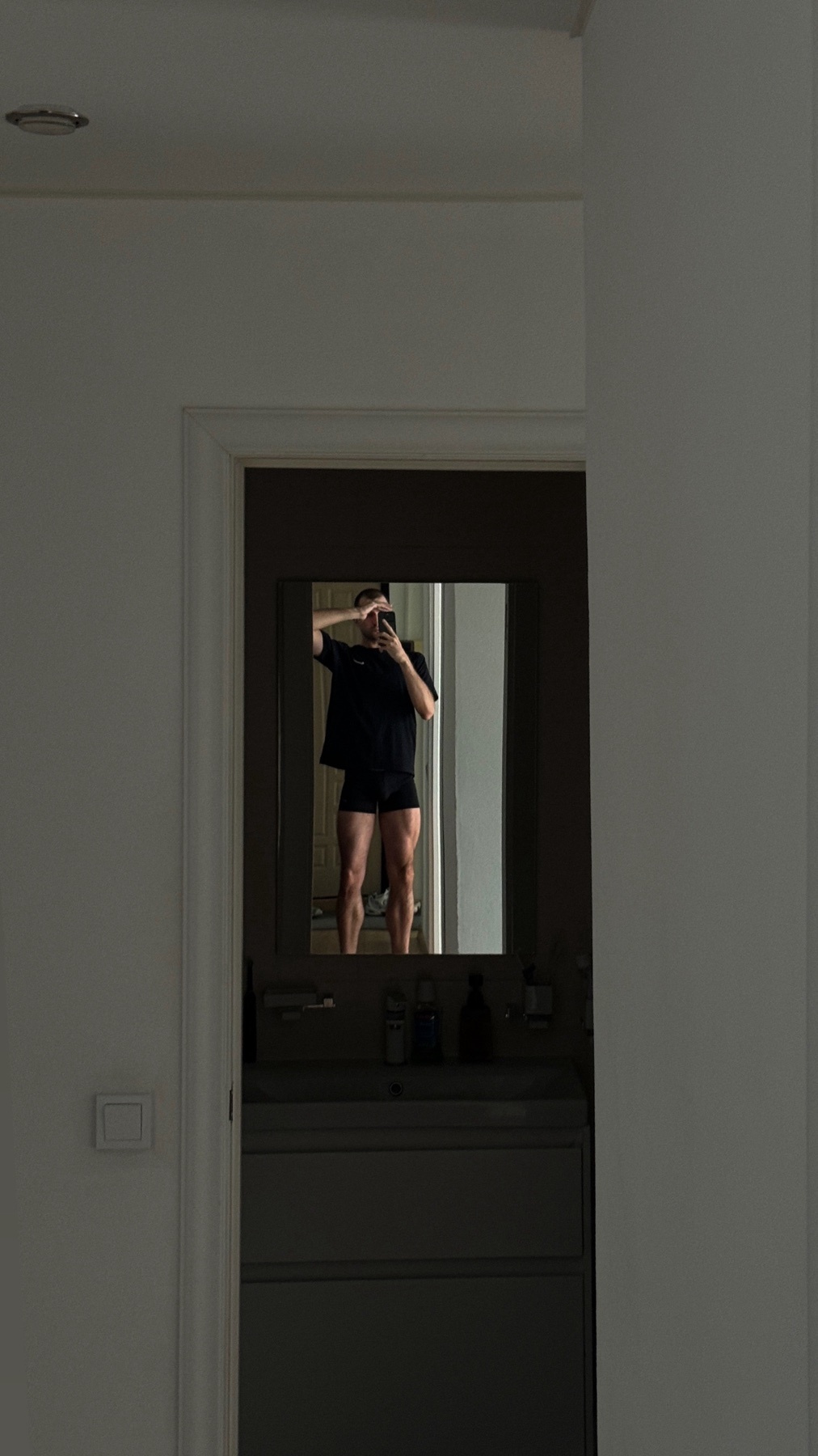 A man takes a mirror selfie in a bathroom, wearing a black t-shirt and shorts. His muscular legs are visible in the reflection.