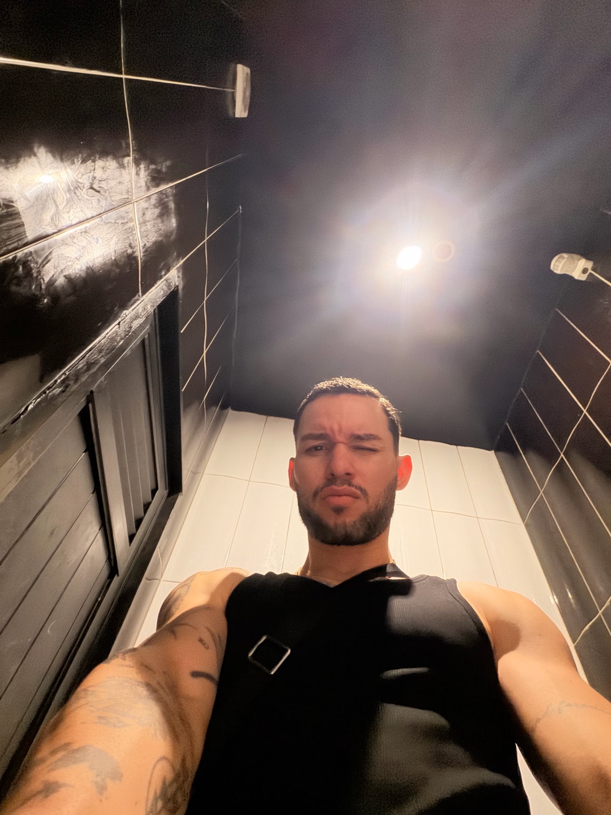 A man takes a selfie in a bathroom. He is shirtless and flexing his arm.
