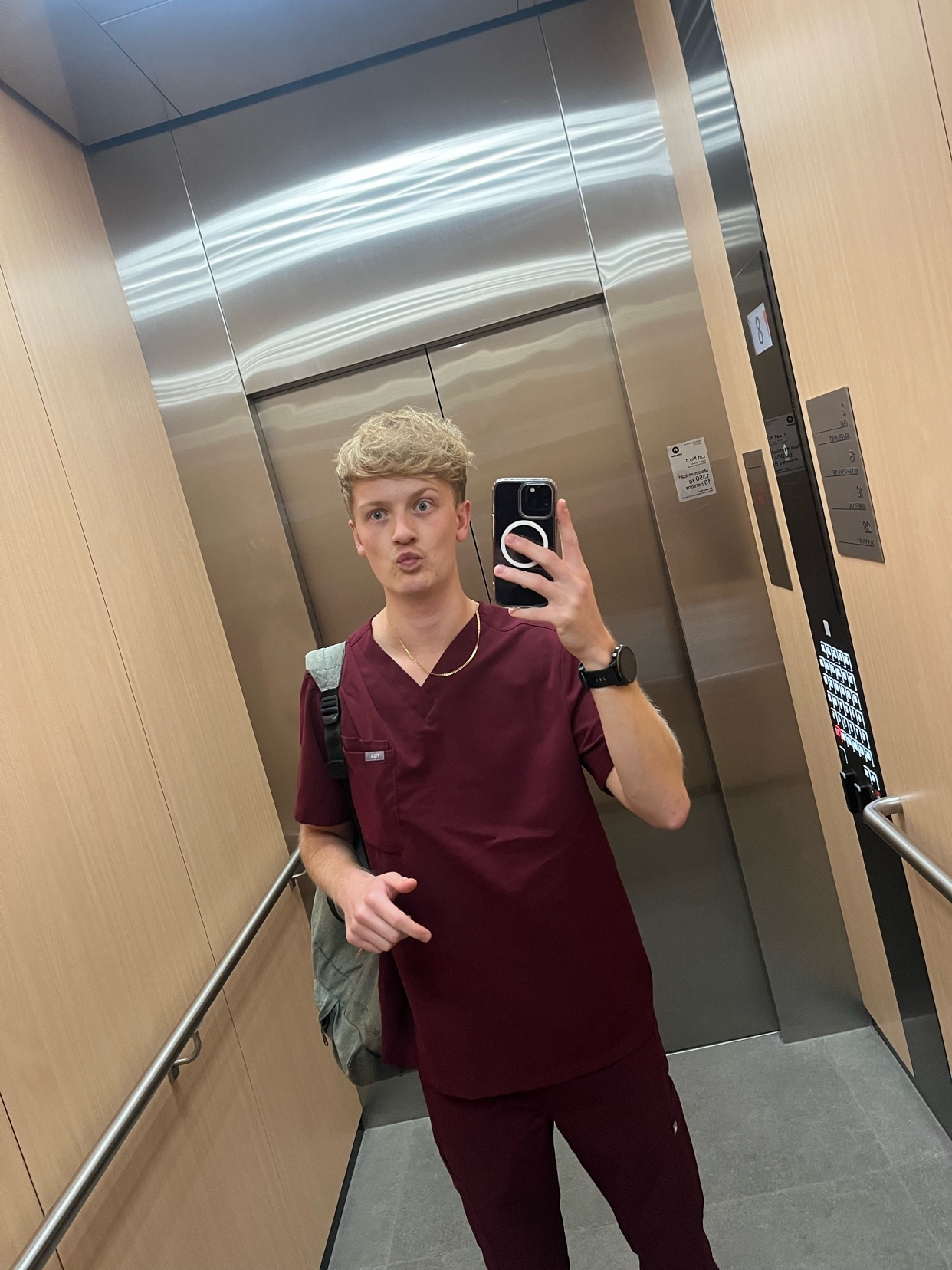 A young man in maroon scrubs poses for a selfie in an elevator. He has short, blond, curly hair and is wearing a gold chain.