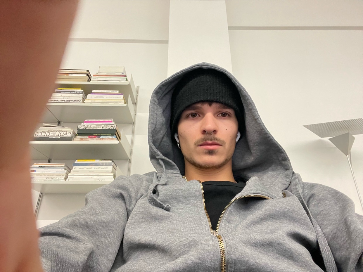 A man in a gray hoodie and black beanie takes a selfie in an office setting. He appears tired.