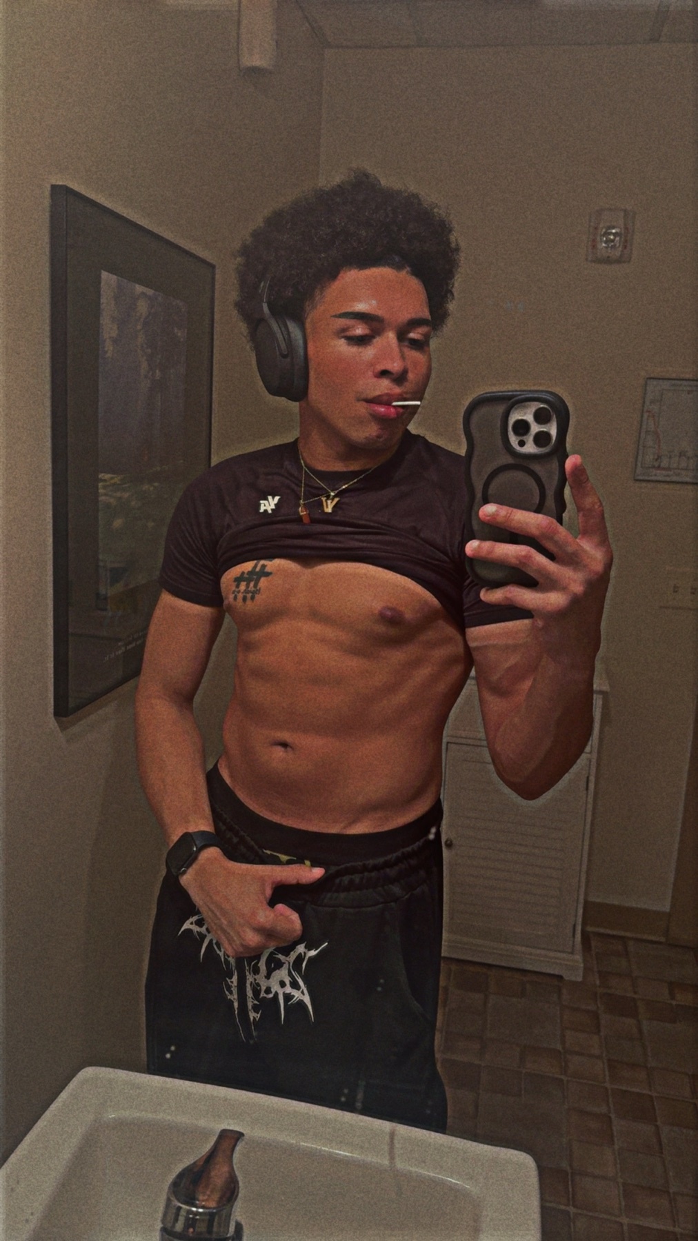A young man with curly hair and headphones takes a mirror selfie while holding a lollipop and wearing a t-shirt pulled up to reveal his abs. He is wearing black pants with white lettering.